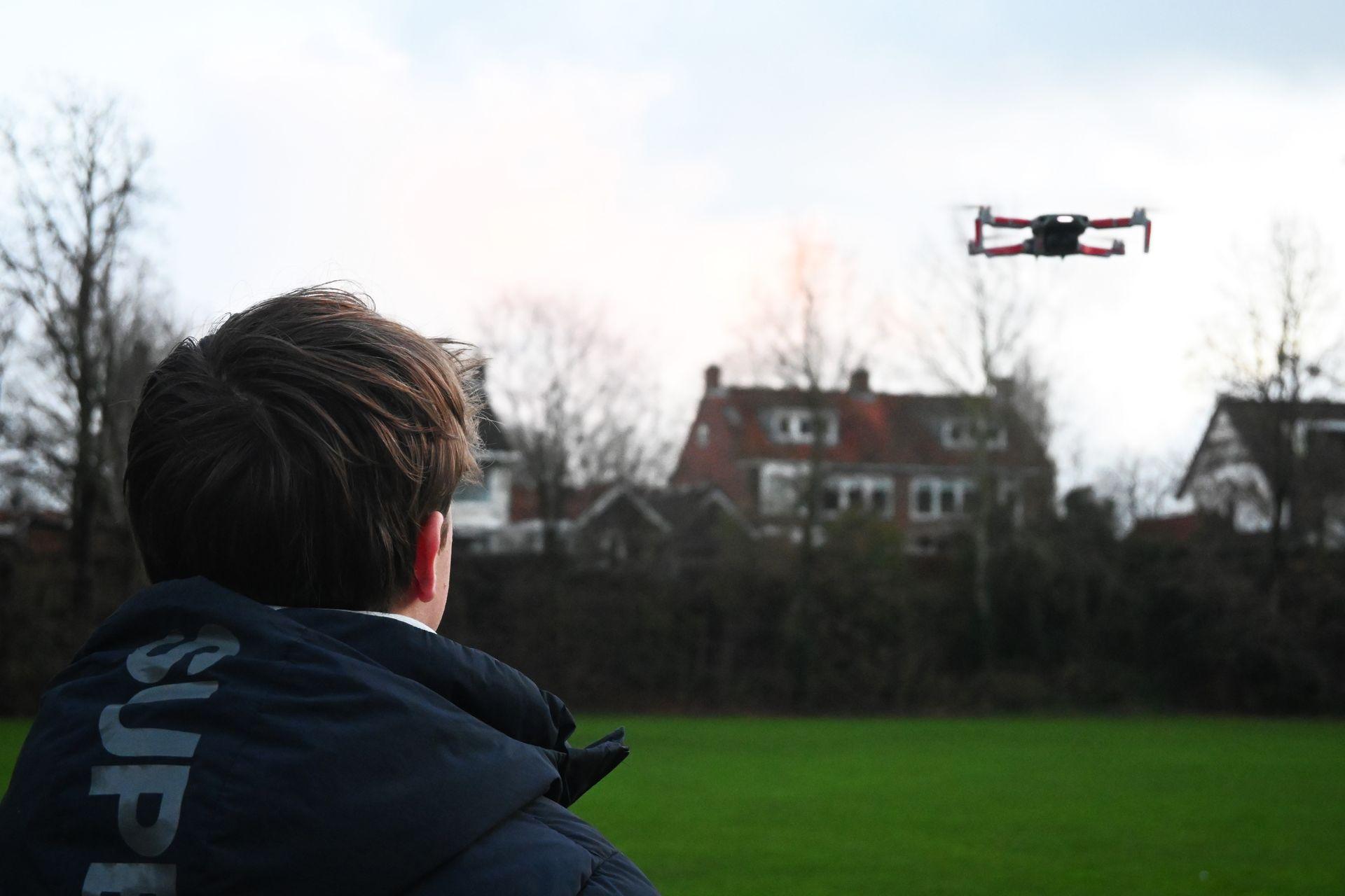 Person watching a flying drone in a grassy field with houses and leafless trees in the background.