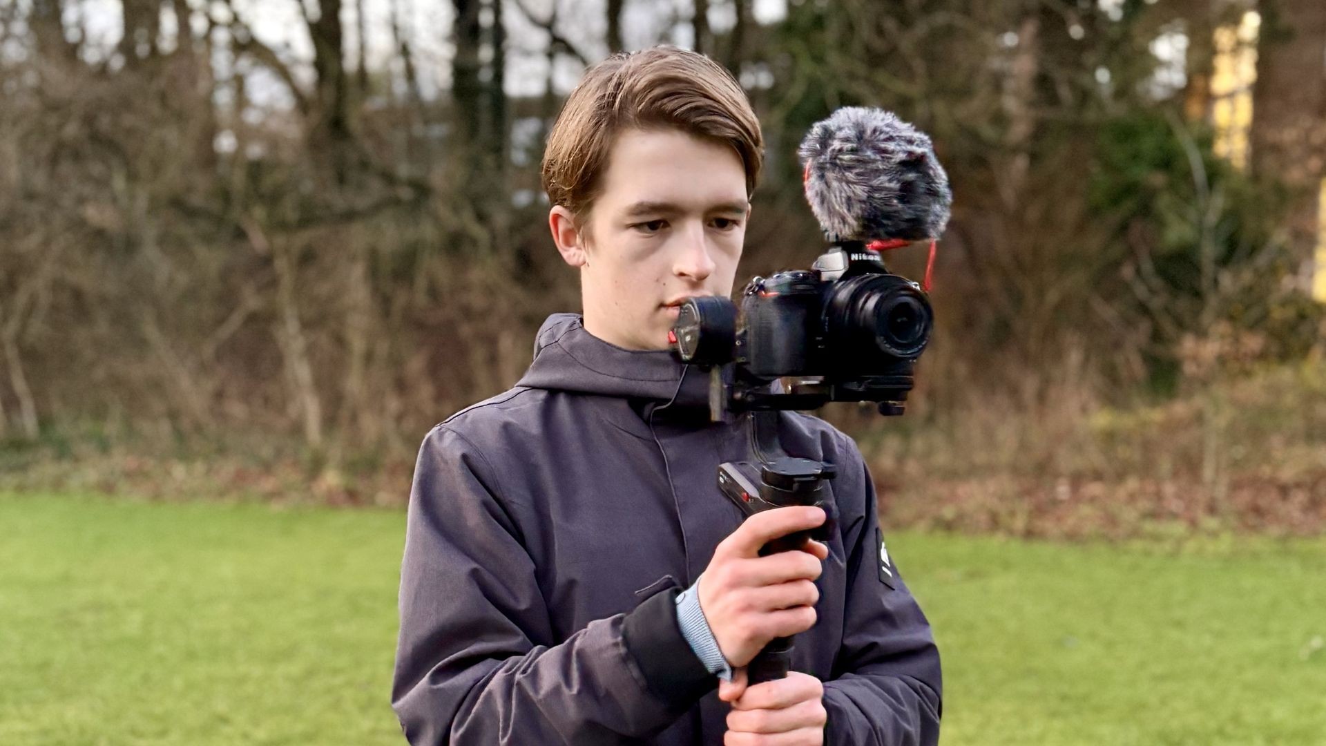 Person holding a camera with a mounted microphone on a gimbal in an outdoor park setting.