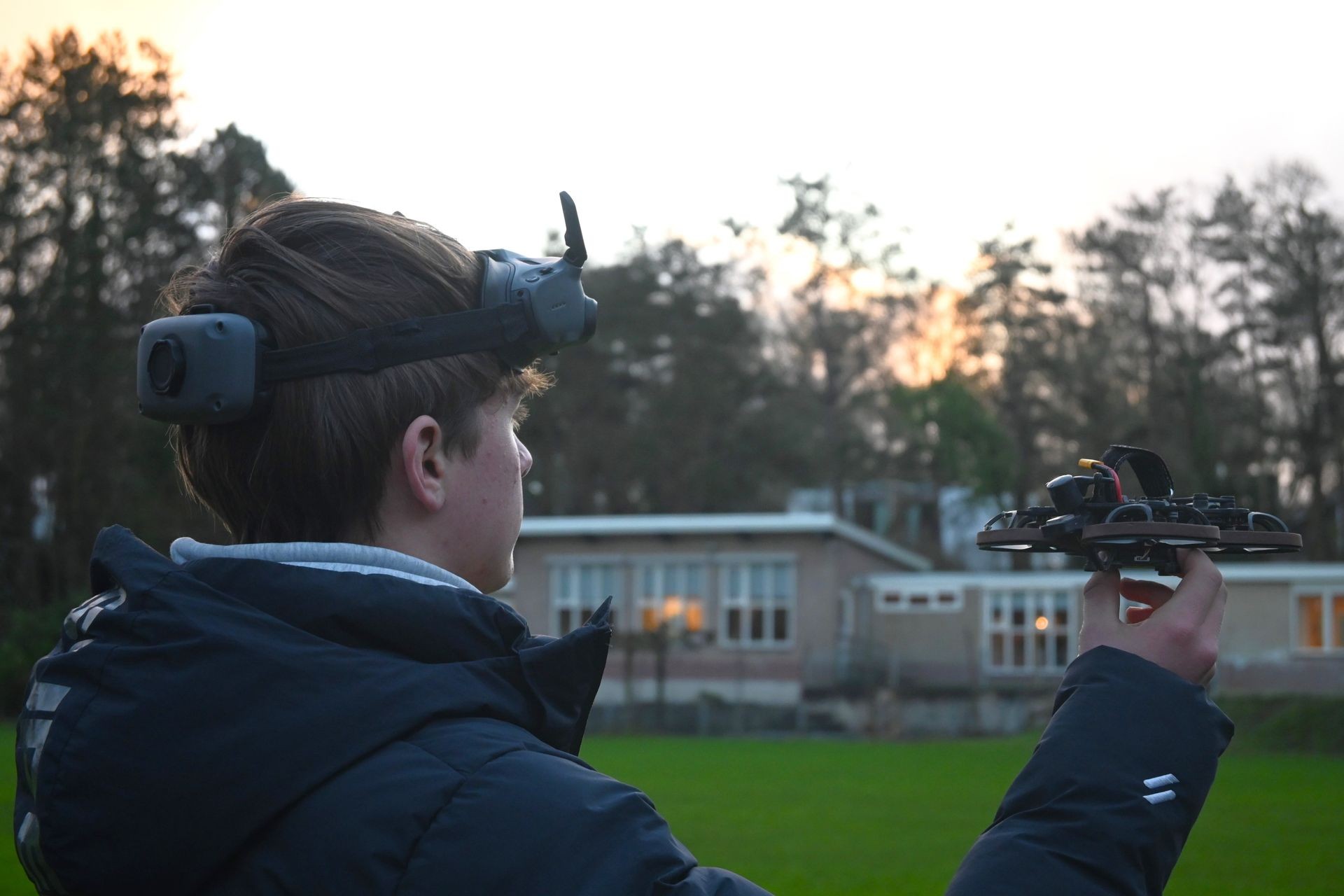 Person wearing VR goggles holding a drone outdoors near a building at dusk.